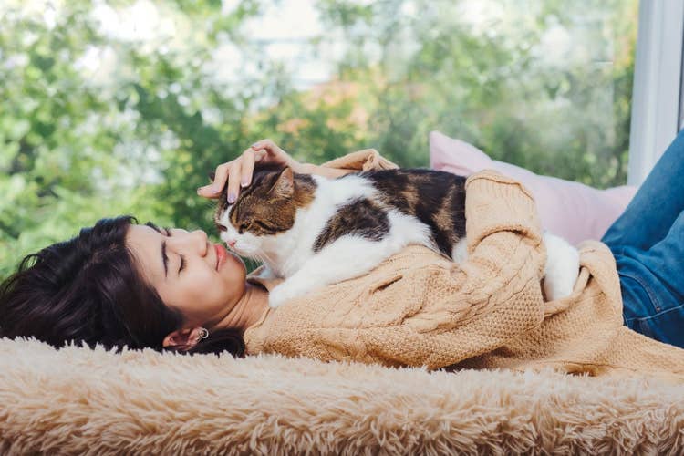 A female owner with dark hair lying down in a window seat with her tortoiseshell and white cat snuggled on her chest.