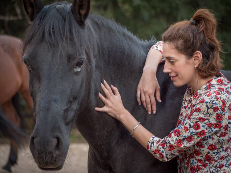 A woman wearing a bright floral shirt awestruck as she calmly strokes a black horse's neck.